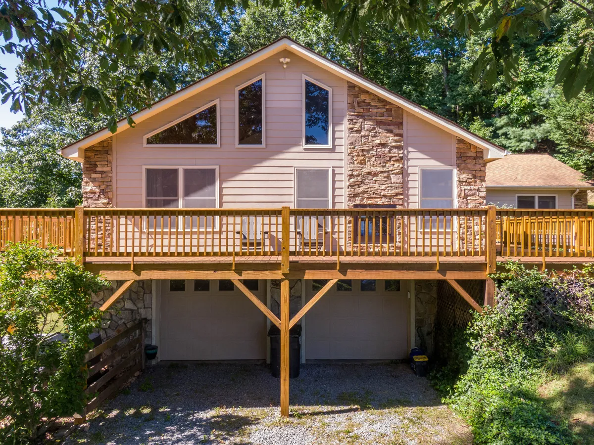 The image captures the home's wooden deck with a sturdy stone facade and large windows, all nestled amidst tall trees. The deck offers an inviting space for relaxing, soaking in the natural setting, and enjoying sunrise or sunset views.