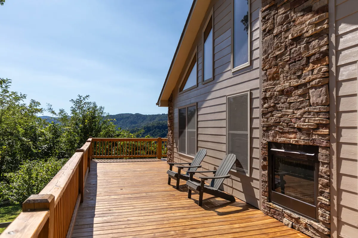 A wooden deck stretches out with adirondack chairs facing a panoramic mountain view. The deck is bordered by a rustic rock façade and surrounded by lush greenery, capturing the transition from the cozy indoor spaces to the expansive outdoors. The scene suggests opportunities for both relaxation and social gatherings, with ample space for outdoor activities.