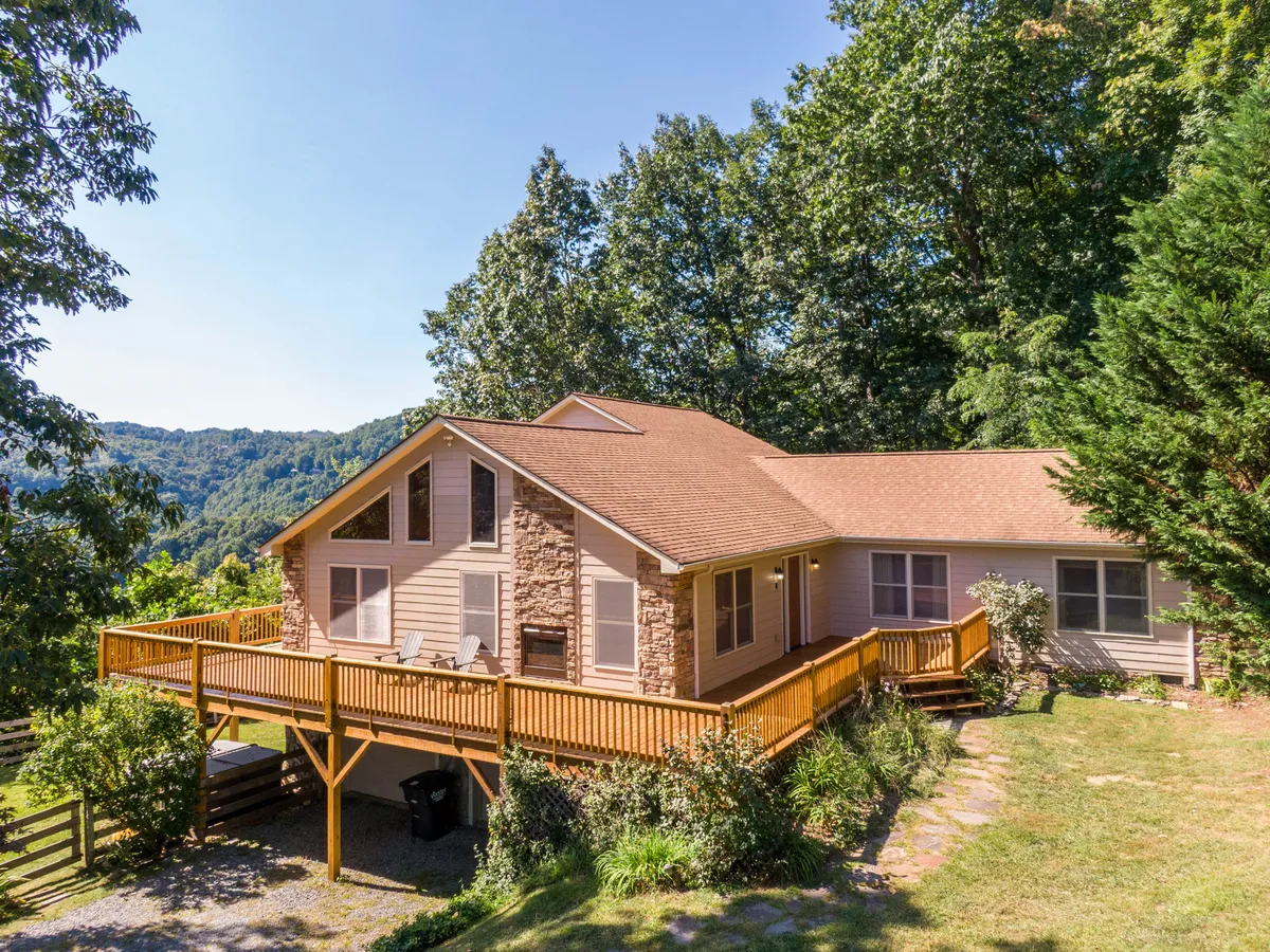 A tranquil exterior view of the home shows a large wooden deck surrounded by lush greenery and tall trees. Expansive windows invite natural light, while the stone facade adds rustic mountain charm. The deck provides ample space for outdoor relaxation or socializing, ideal for humans and pets alike.