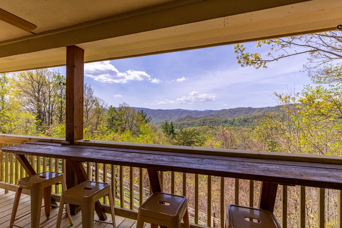 A cozy outdoor bar counter with stools overlooks a sweeping mountain vista. The scene captures a sunrise or sunset moment, evoking a sense of relaxation and camaraderie. The open-air setup is perfect for sipping drinks, sharing stories, and enjoying nature's beauty with friends.