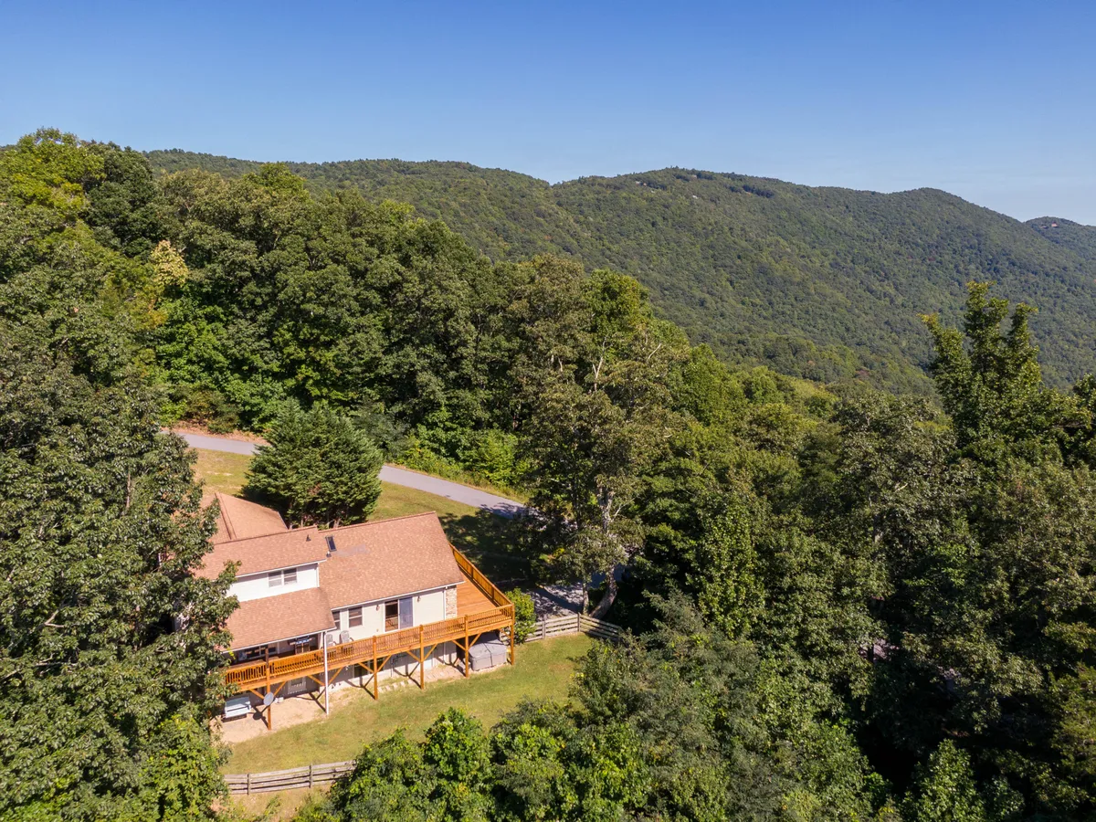 A wide exterior view of the house nestled among rolling hills and mountains, with a generous wooden deck offering panoramic vistas of the surrounding landscape. The home's setting is peaceful and expansive, with open grassy areas ideal for family activities.