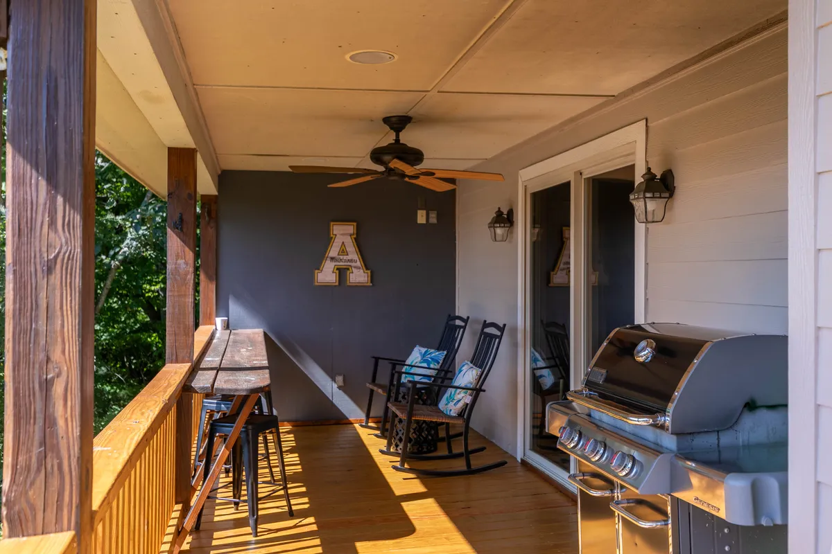 A cozy outdoor area features rocking chairs, a grill, and a bar table on a wooden porch. The setting is casual and inviting, surrounded by greenery—perfect for socializing, barbecuing, or relaxing with pets after a day of local exploration.