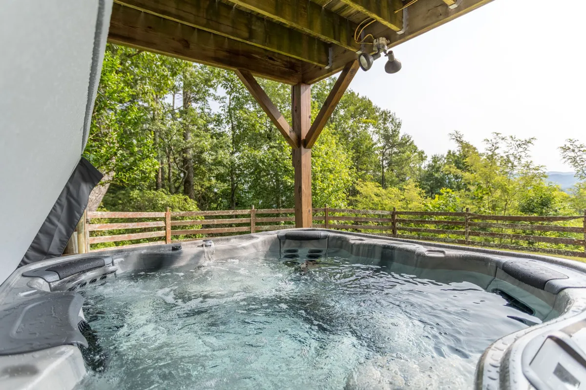 A tranquil scene featuring a spacious hot tub nestled on the deck, framed by sturdy wooden beams and surrounded by nature. The bubbling water invites relaxation, while the open backdrop showcases rolling hills fading into the sunset.