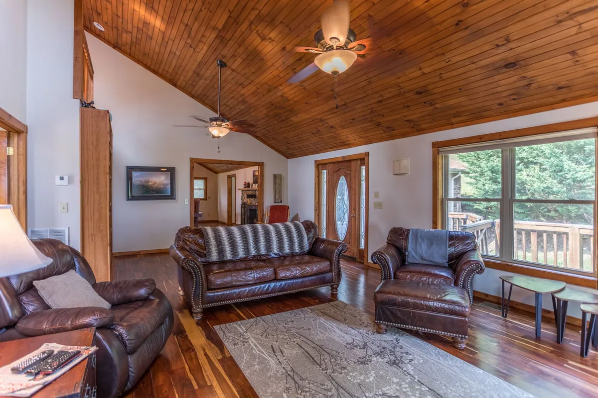 A living area with leather sofas, a wooden ceiling, a colorful rug, and a large window overlooking the outdoors. The mood is welcoming, perfect for relaxing, socializing, or reading by the window.