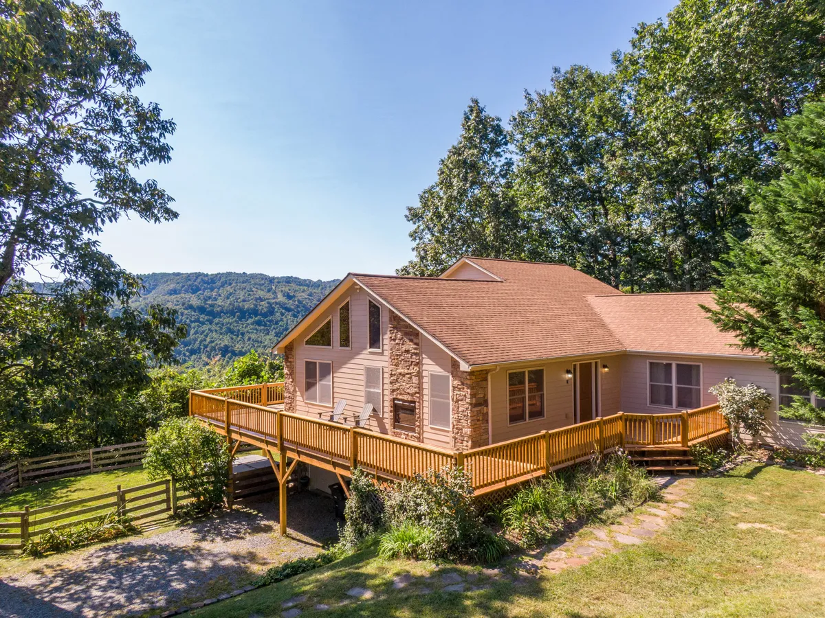 The image showcases a spacious single-family mountain home with a welcoming wooden deck, nestled among tall trees and overlooking sweeping Blue Ridge mountain vistas. The setting radiates privacy and tranquility, with the deck positioned perfectly for sunset views and gatherings. The lush forest and distant peaks invite exploration and relaxation, hinting at the property's secluded five-acre grounds.