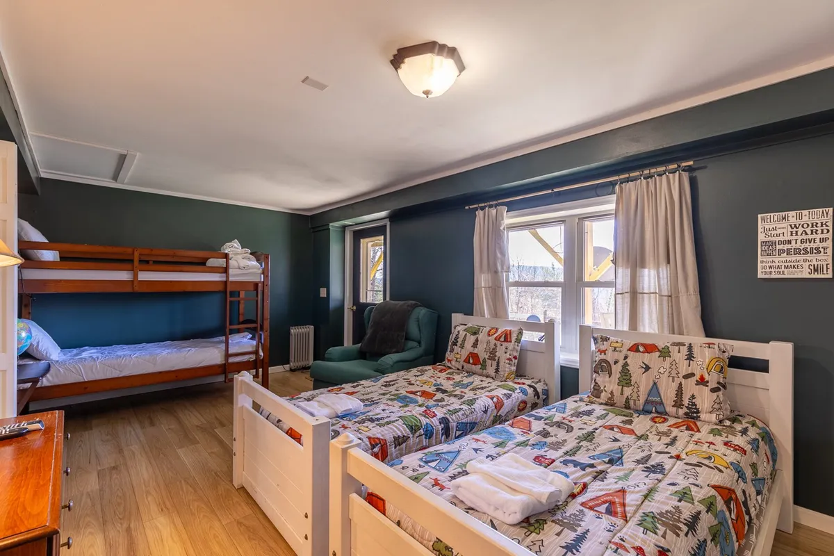 A playful and welcoming bedroom featuring a bunk bed and twin beds, a cozy chair, and a large window offering a view. The decor is colorful and family-friendly, making it ideal for children or groups of friends.
