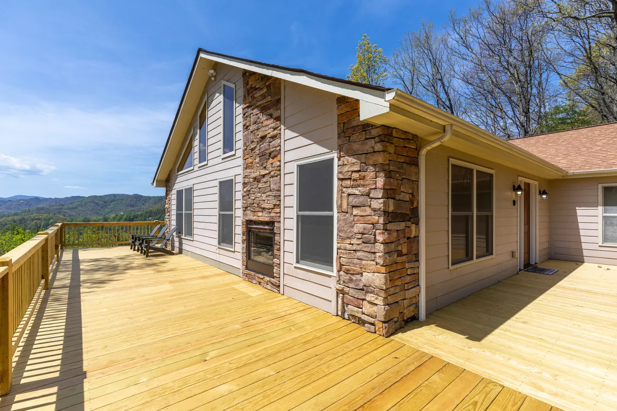 A wooden deck with a stone facade and large windows, offering unobstructed views of the mountains beyond. Adirondack chairs suggest a relaxing outdoor dining or lounging area, with the sun setting over the hills.