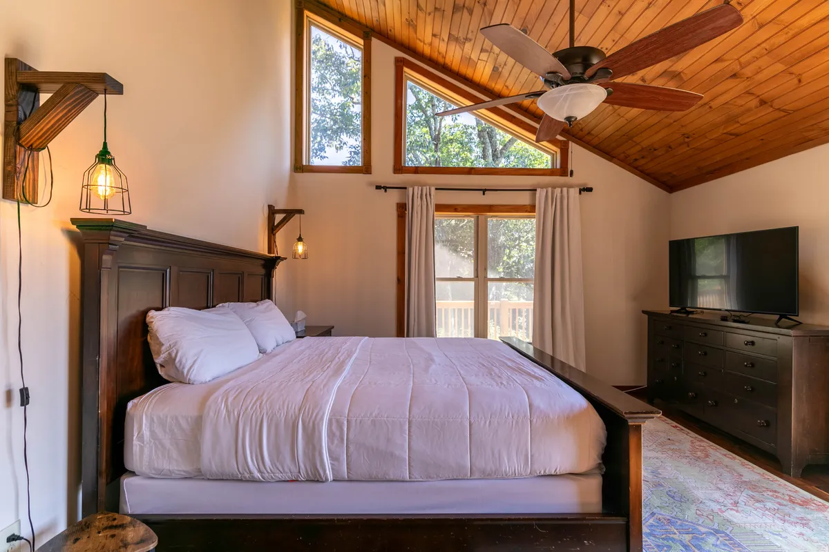 A spacious bedroom featuring a large bed with plush bedding, wooden furniture, a ceiling fan, and expansive windows letting in abundant natural light. There's a television positioned for comfortable viewing and cozy corners ideal for reading or relaxing.
