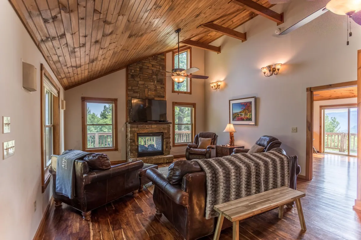 The image showcases a warm, inviting living room with a striking stone fireplace as the centerpiece. Leather sofas are arranged for intimate conversation or group gatherings, while large windows frame scenic outdoor views. The wooden ceiling adds rustic charm and enhances the cozy mountain retreat atmosphere.